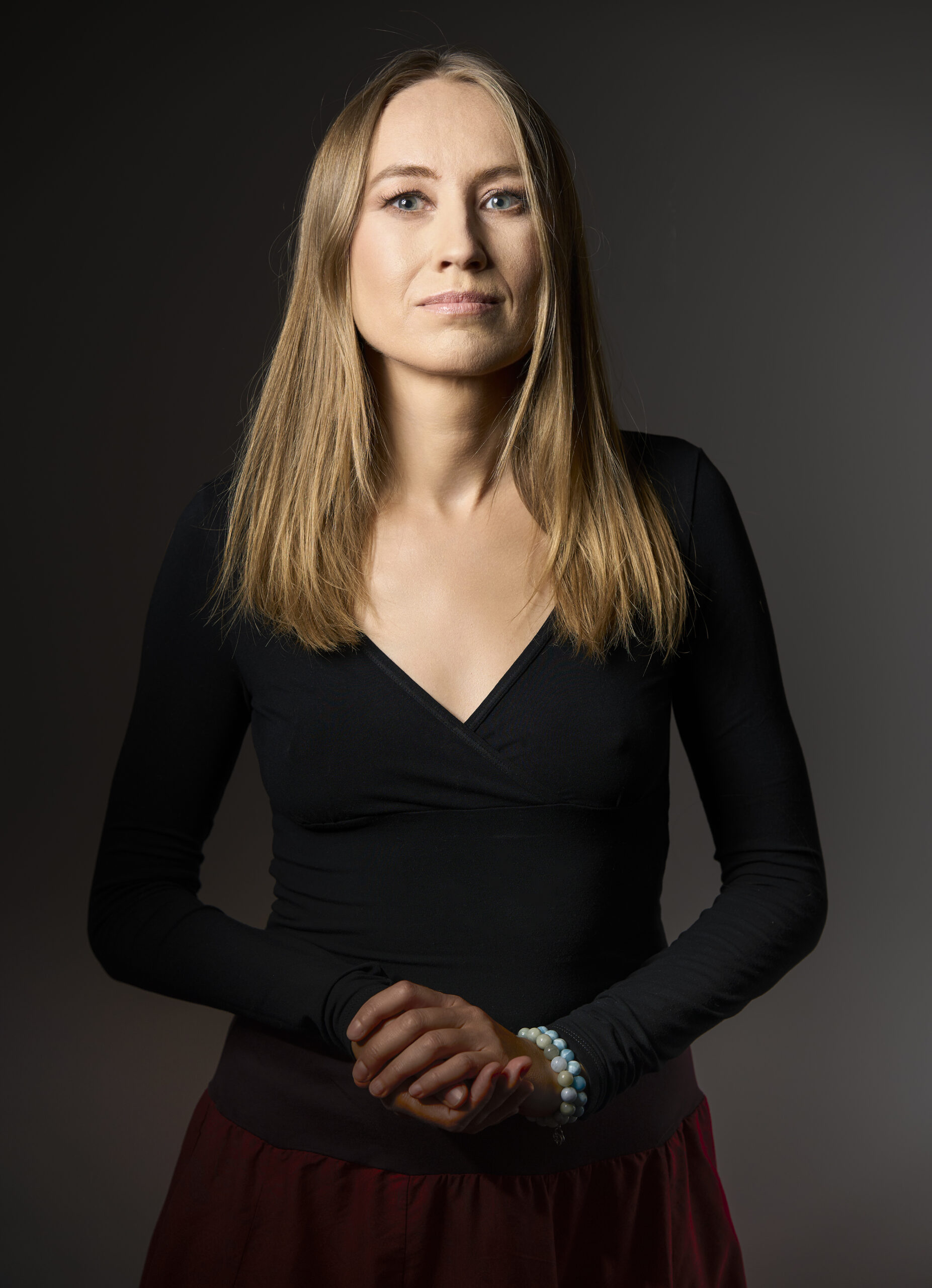 Portret studyjny Małgorzaty Żabińskiej - — blond włosy, ubrana w czarną bluzkę, dłonie splecione przed sobą, ciemne tło. Terapeutka o wysokich kompetencjach i wieloletniej praktyce w pracy z ciałem.