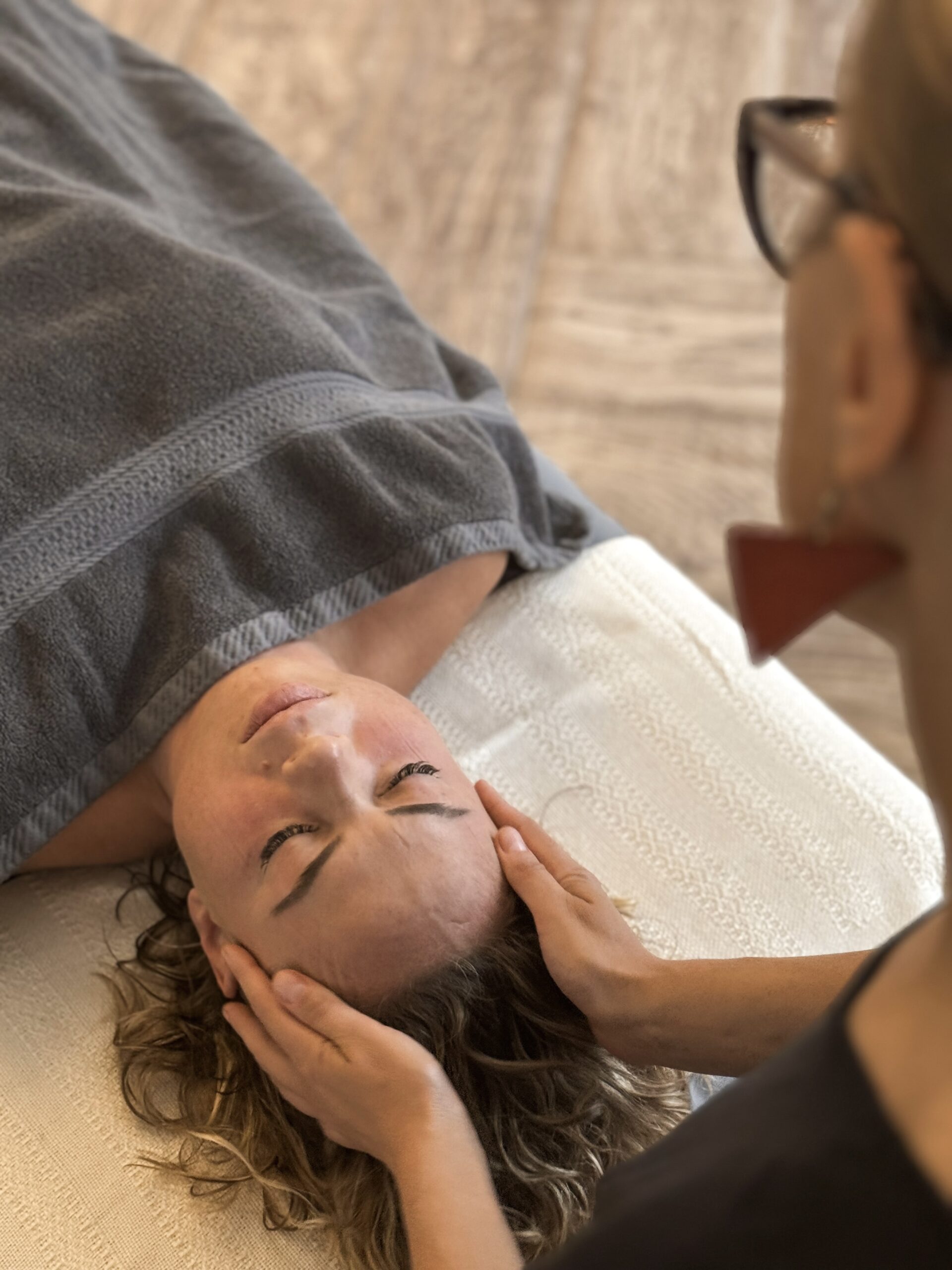 Terapeutka Małgorzata Żabińska obejmuje swoimi rękoma głowę kobiety leżącej na stole do masażu podczas wykonywania Masażu Relaksacyjnego. Osoba przyjmująca masaż jest przykryta szarym ręcznikiem.